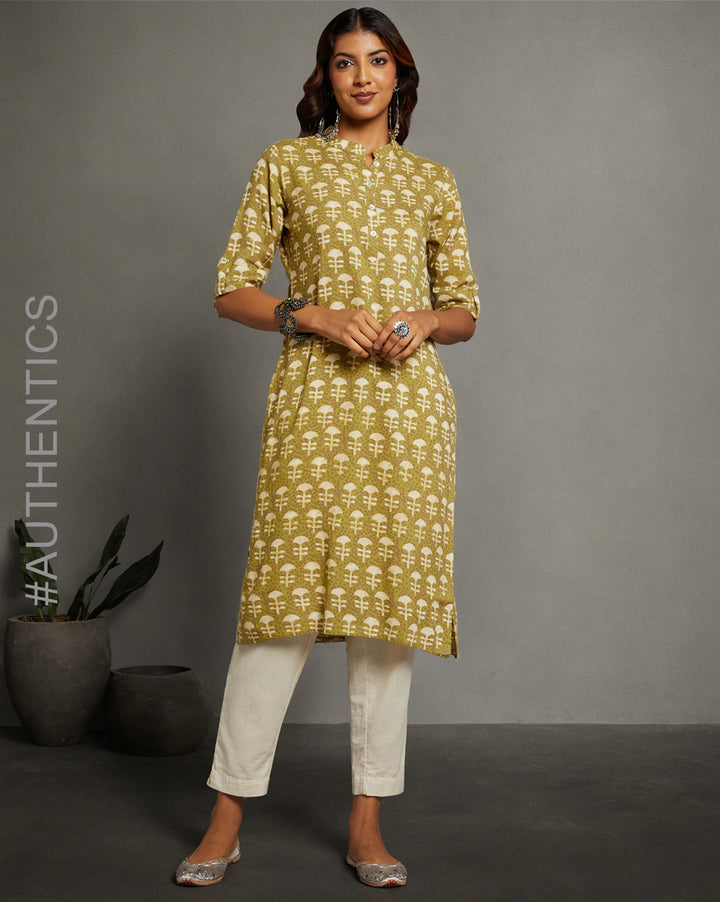 Stylish Printed green Cotton dabu print Kurti with three-fourth sleeves paired with white pant, worn by a woman standing next to a flower pot with open hair.
