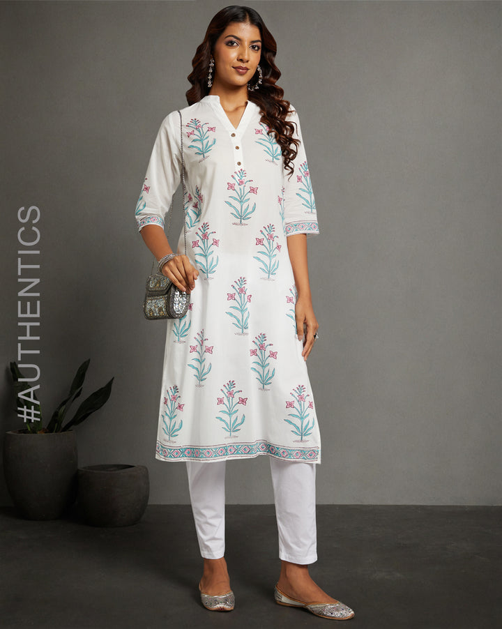 Stylish white Kurti with jaipuri floral print and three-fourth sleeves paired with white pant, worn by a woman standing next to a flower pot with open hair.
