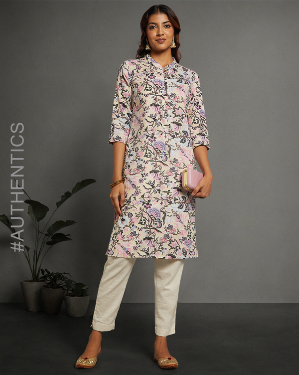 Stylish Pastel Yellow Floral Hand Block Cotton V-Neck Kurta, paired with white pant and jutti, worn by a woman standing next to flowerpot.
