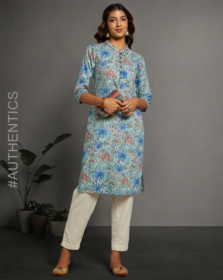 Stylish Sky blue floral printed kurta knee length and three-fourth sleeves paired with white pant and golden jutti, worn by a woman standing next to flowerpot.