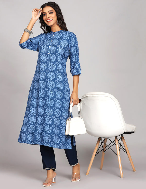 Stylish Blue Long Kurti with Collar Neck Line, paired with navy blue pant and white transparent sandals, worn by a woman standing next to chair.