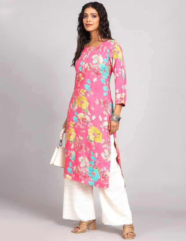 Stylish  Pink Color Floral Print Long Kurti with Round Neck design with white wide palazzo and brown transparent sandals, worn by a woman standing in white background.
