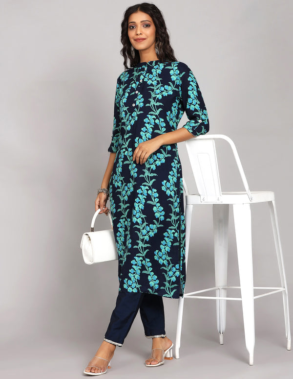 Stylish Navy Blue Floral Printed Long Kurti with Collar Neck Line with blue pant and white transparent sandals, worn by a woman standing next to chair.