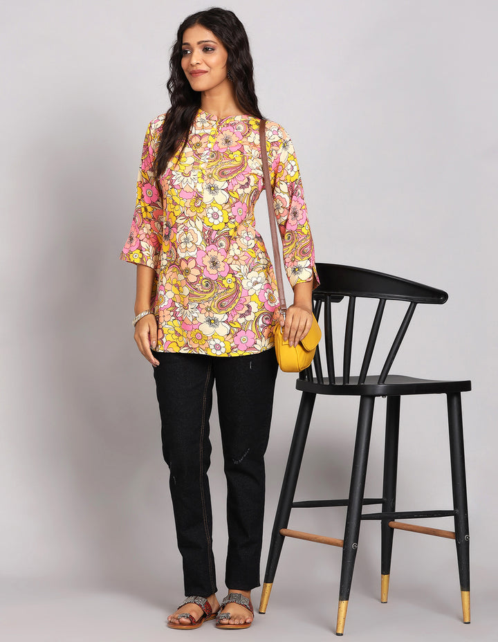 Stylish Multi Color Short Kurti with Floral Print,paired with black jeans and brown slippers, worn by a woman standing next to a chair.