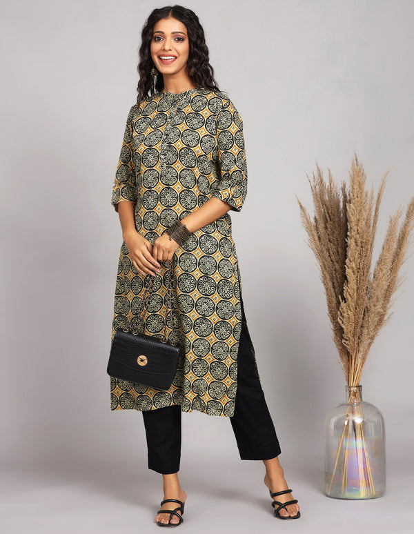 Stylish  Rust Yellow Kurti with hand block Print, paired with black pant and black heels, worn by a woman standing next to flower pot with open hair.
