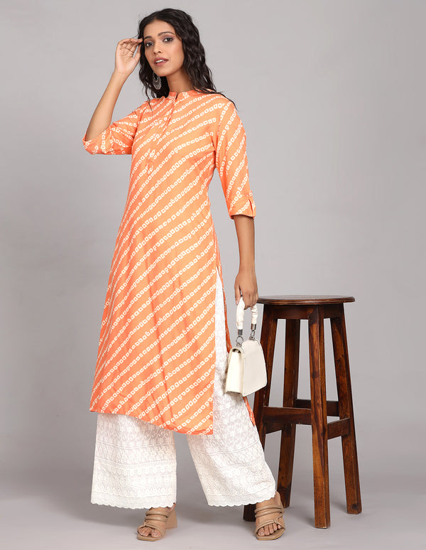 Stylish orange color printed long kurti, paired with white wide leg palaazo and brown sandals, worn by a woman standing next to table.