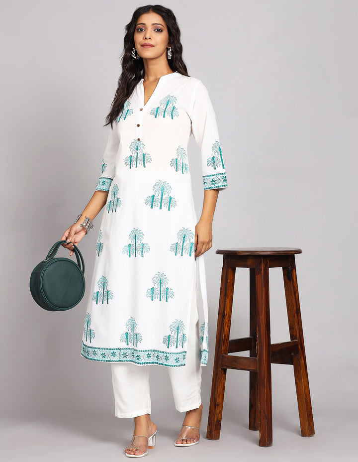 Stylish White Long Kurti With Hand Block Print with white pant and white transparent sandals, worn by a woman standing next to table.