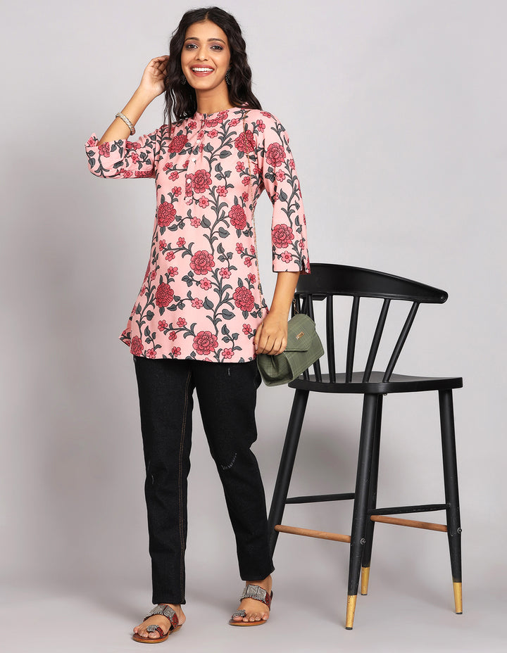 Stylish Chalk Pink floral printed Short Kurti, paired with black jeans and brown slippers, worn by a woman standing next to chair.