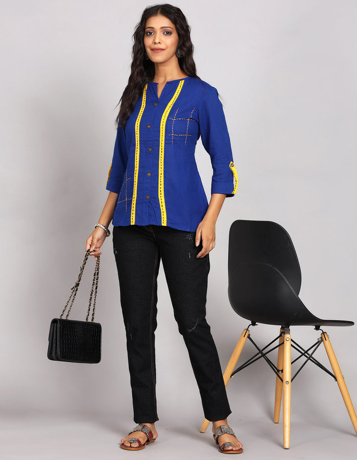 Stylish Royal blue top with yellow strips and wooden buttons, paired with fitted black jeans and brown flat sandals, worn by a woman standing next to a chair.