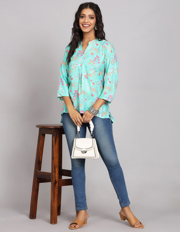 Stylish Aqua Blue Floral Print Short Kurti,paired with blue skiny jeans and brown sandals, worn by a woman standing next to a stool taking a white handbag in hands.