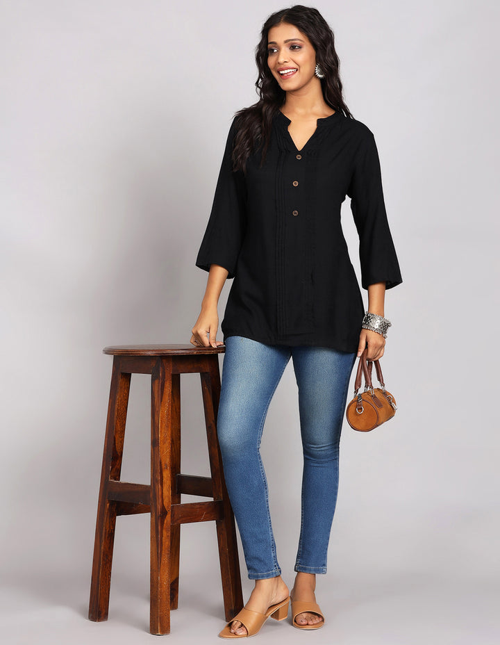 Stylish black top with wooden buttons, paired with fitted blue jeans and brown heeled sandals, worn by a woman standing next to a stool.